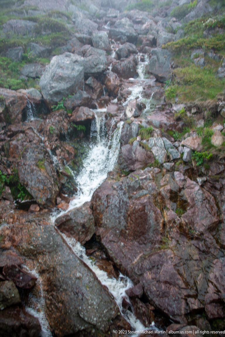 Narrow stream cascading over reddish exposed bedrock with sparse low green vegetation along the edges.