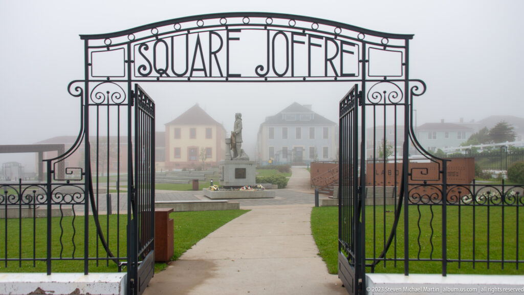Open wrought-iron gates reading LE SQUARE JOFFRE frame a path, central monument and buildings in fog.