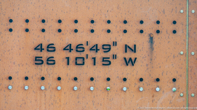 Rust-colored perforated metal panel cut with the coordinates 46°46′49″ N and 56°10′15″ W.