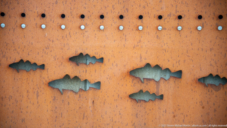 Several fish silhouettes cut through a rust-colored perforated steel panel with blue-gray backing visible through the openings.