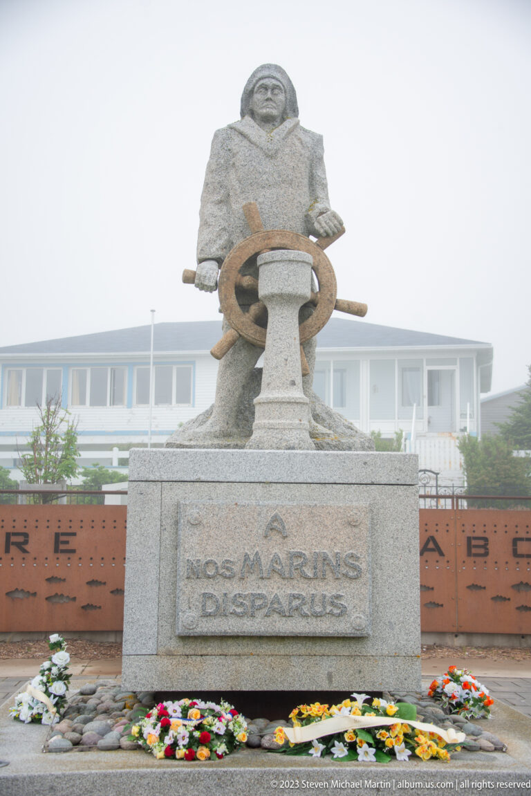Stone memorial of a mariner holding a ship’s wheel above the inscription NOS MARINS DISPARUS, with flowers at the base.