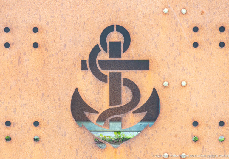 Stylized anchor emblem on a rust-colored perforated metal panel, with a small opening revealing greenery below.