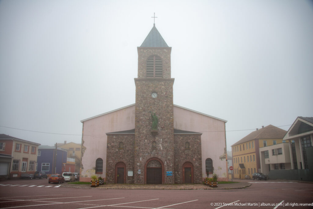 Front view of a stone church with square bell tower, clock, arched doors and cross, surrounded by foggy town buildings.