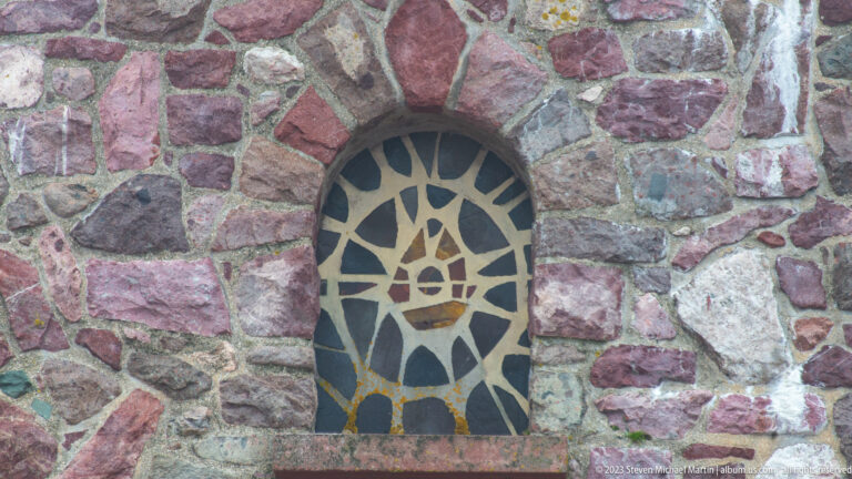 Arched mosaic with a stylized boat-like emblem and radiating dark-and-cream pattern set into a multicolored stone wall.