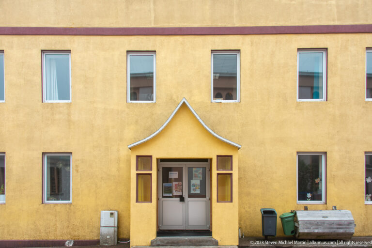 Yellow two-story building with regularly spaced windows and a small pointed canopy above the central double doors.