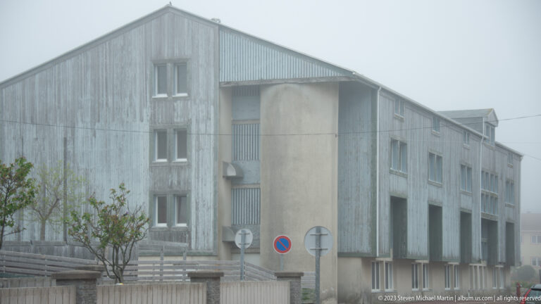 Large gray concrete building with repetitive vertical bays seen through dense fog behind roadside signs and a low wall.