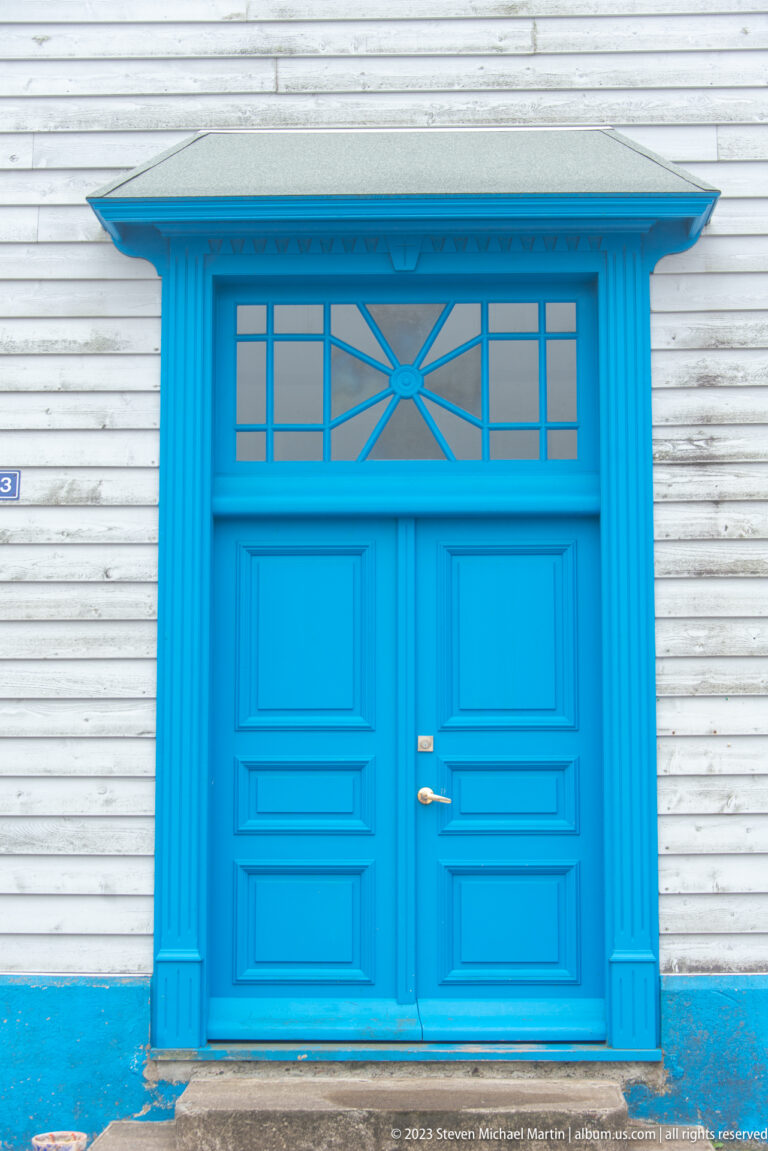 Bright blue paneled double doors with matching trim and a geometric glazed transom set in white horizontal siding.