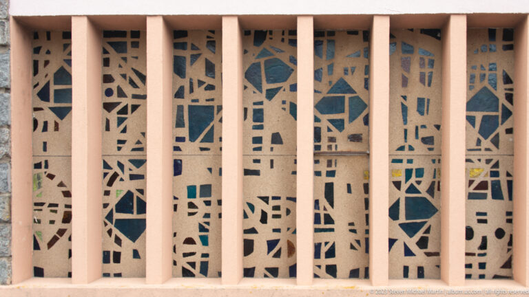 Abstract perforated concrete screen with irregular openings seen behind evenly spaced pale pink vertical supports.