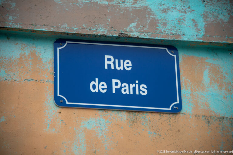 Blue “Rue de Paris” street sign mounted on a weathered turquoise-and-ochre wall.