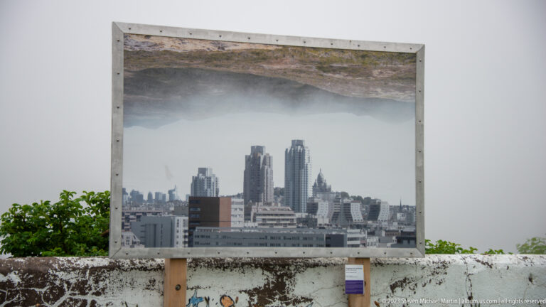 Large framed city-skyline photograph mounted outdoors against a foggy background with low vegetation and a wall.