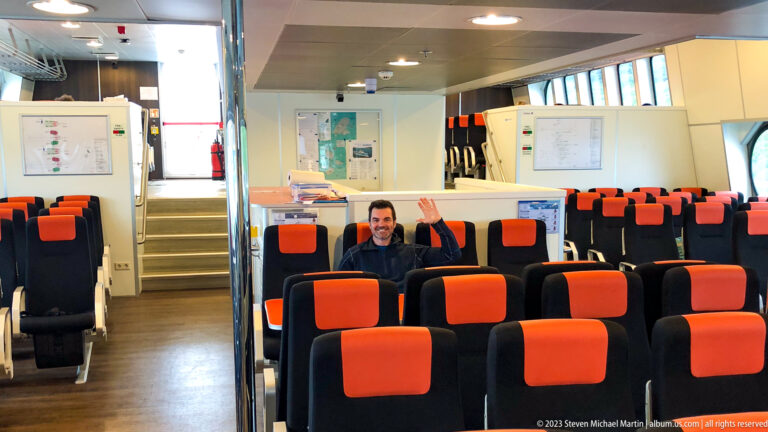 Rows of black-and-orange ferry seats with a seated passenger waving from the center of the cabin.