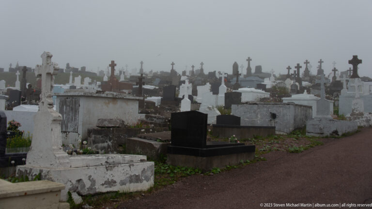 Foggy cemetery filled with closely spaced crosses, stone tombs, family monuments, and a reddish walking path.