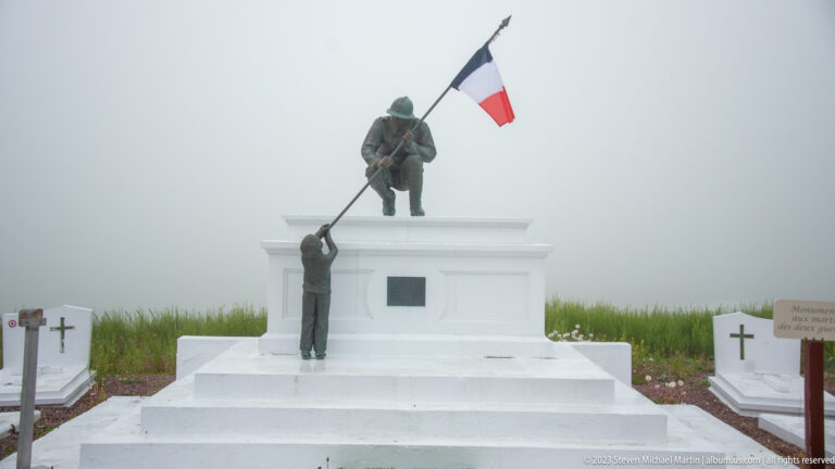 White cemetery memorial with two bronze figures, one holding a French tricolor flag aloft in dense fog.