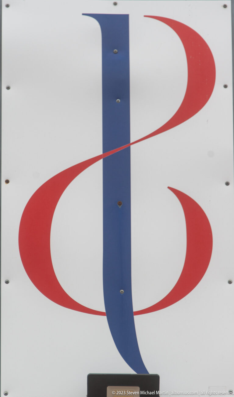 Abstract red-and-blue emblem on a white panel, formed by a vertical blue stroke and two curved red elements.