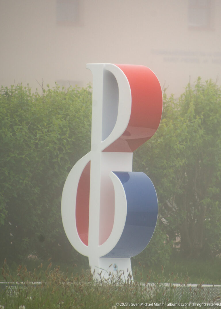 Freestanding red, white, and blue abstract emblem beside a hedge in misty conditions.