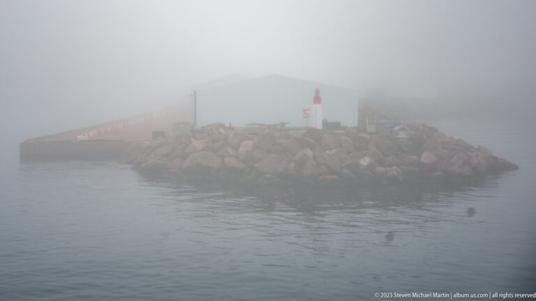 Red-and-white harbor beacon on a boulder breakwater fading into dense fog over calm water.