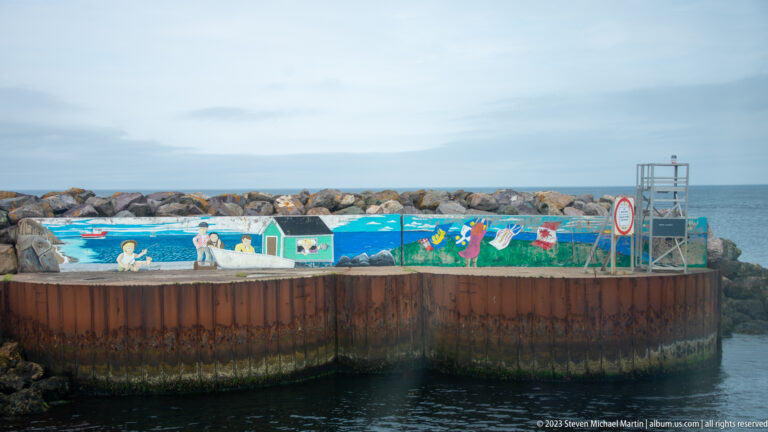 Curved steel harbor wall painted with colorful maritime murals, backed by rocks and open water.