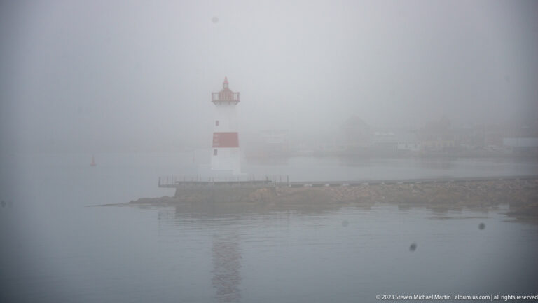Pointe aux Canons lighthouse, white with a red band and lantern, standing on a breakwater in thick fog.
