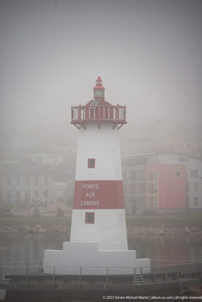 Close view of Pointe aux Canons lighthouse with red band, named lettering, and red-roofed lantern in fog.