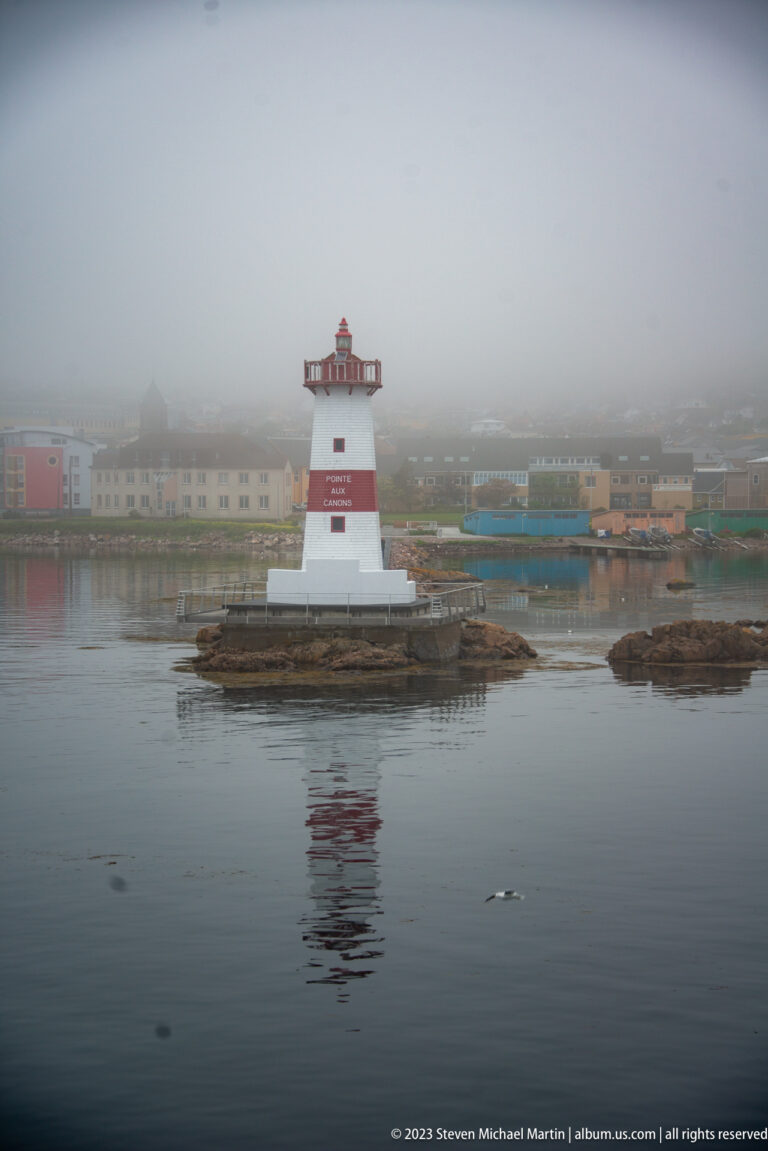 Pointe aux Canons lighthouse on a low rocky islet, with foggy Saint-Pierre waterfront buildings behind it.