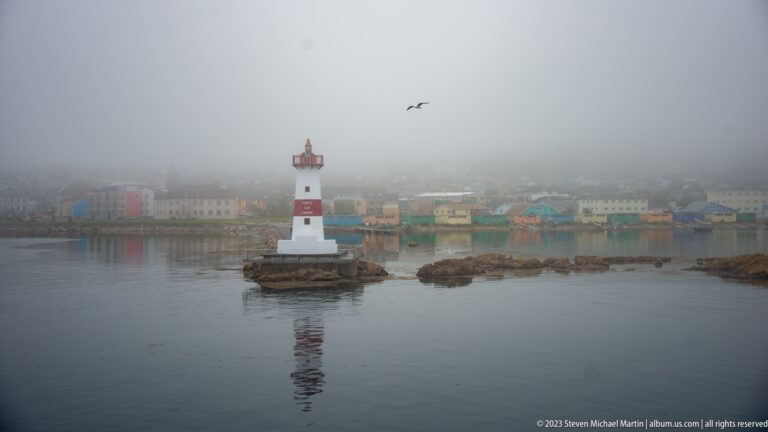 Pointe aux Canons lighthouse reflected in calm water, with colorful fog-softened waterfront buildings and a flying seabird.