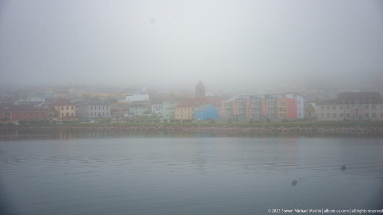 Foggy Saint-Pierre waterfront with pastel buildings and a church tower seen across calm harbor water.