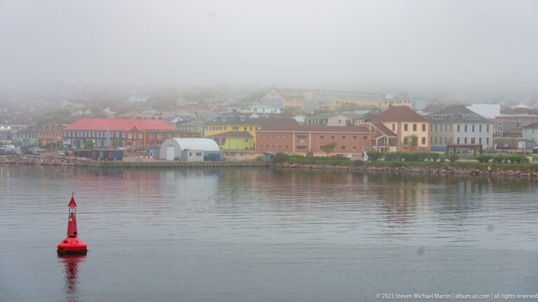 Red navigation buoy floating in calm harbor water before Saint-Pierre’s colorful misty waterfront.