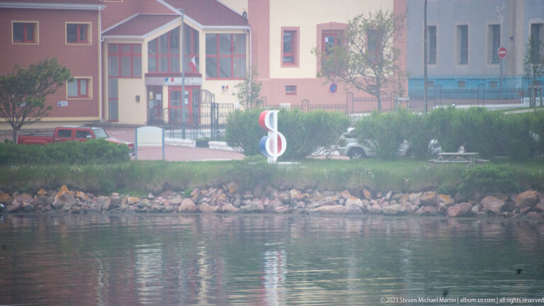 Pastel shoreline buildings, greenery, rocky revetment, and a red-and-white marker beside Saint-Pierre harbor.