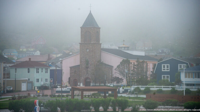 Stone church with a tall square tower and pointed roof rising among houses in foggy Saint-Pierre.