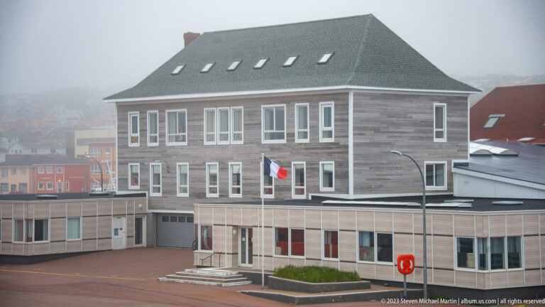 Large gray wood-clad public building with rows of windows, hipped roof, skylights, and a French flag.