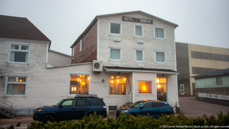 Weathered wood-clad Hôtel Robert building with lit ground-floor windows and parked cars in front.