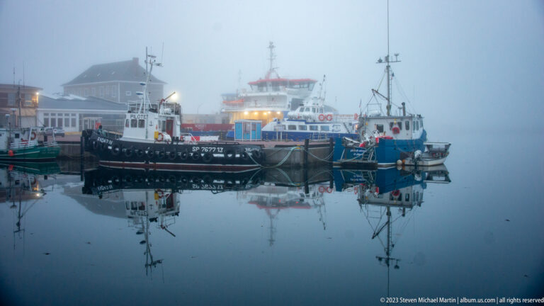 Several fishing and service boats moored together in a foggy harbor, reflected in glassy water.