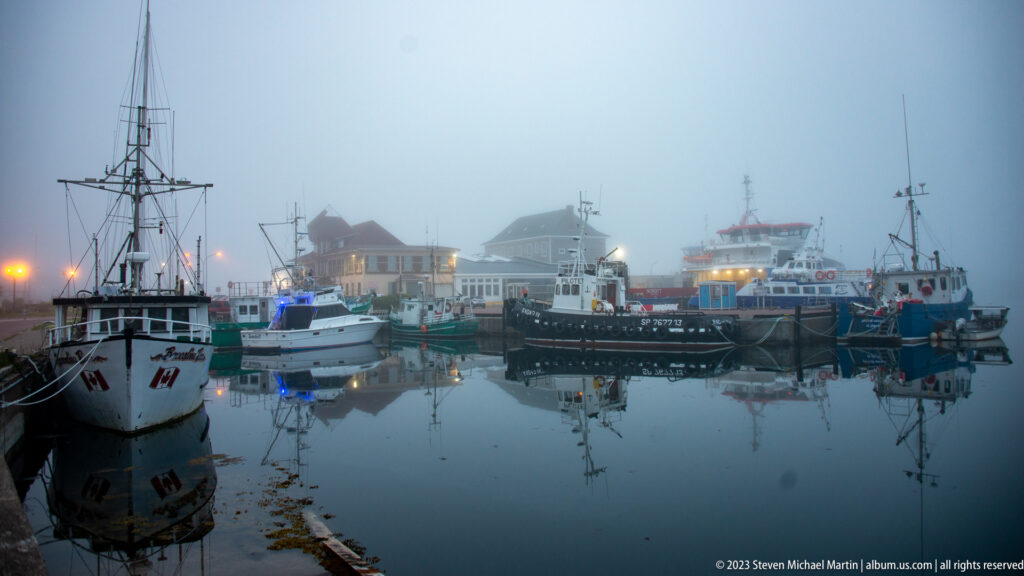 Wide view of fishing boats and work vessels moored in Saint-Pierre harbor under fog, with lights reflected in calm water.