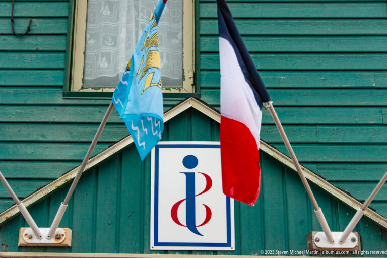 French tricolour and blue territorial flag mounted on a green wooden façade above a Saint Pierre & Miquelon emblem.
