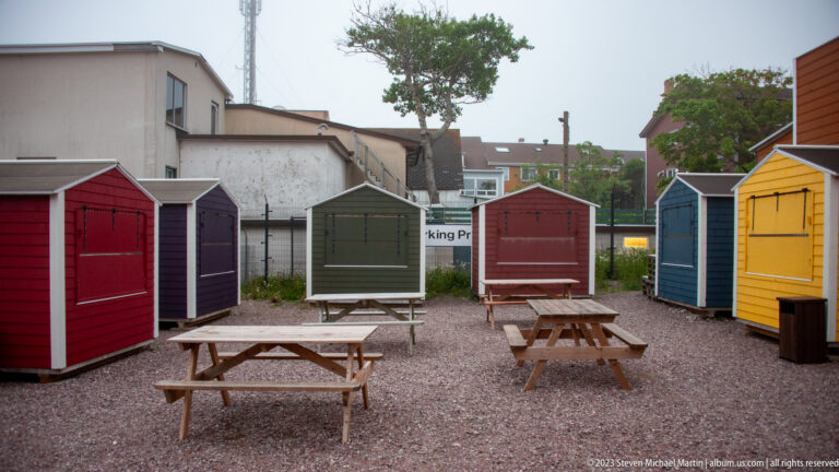 Rows of small red, purple, green, blue, and yellow wooden sheds surrounding picnic tables on gravel.