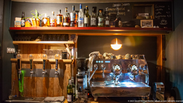 Wood-paneled café bar with beer taps, bottles, glassware, espresso machine, and handwritten chalkboard menus.