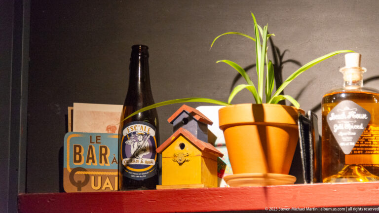 Bar shelf with potted plant, miniature birdhouses, bottles, and a decorative Le Bar du Quai sign.