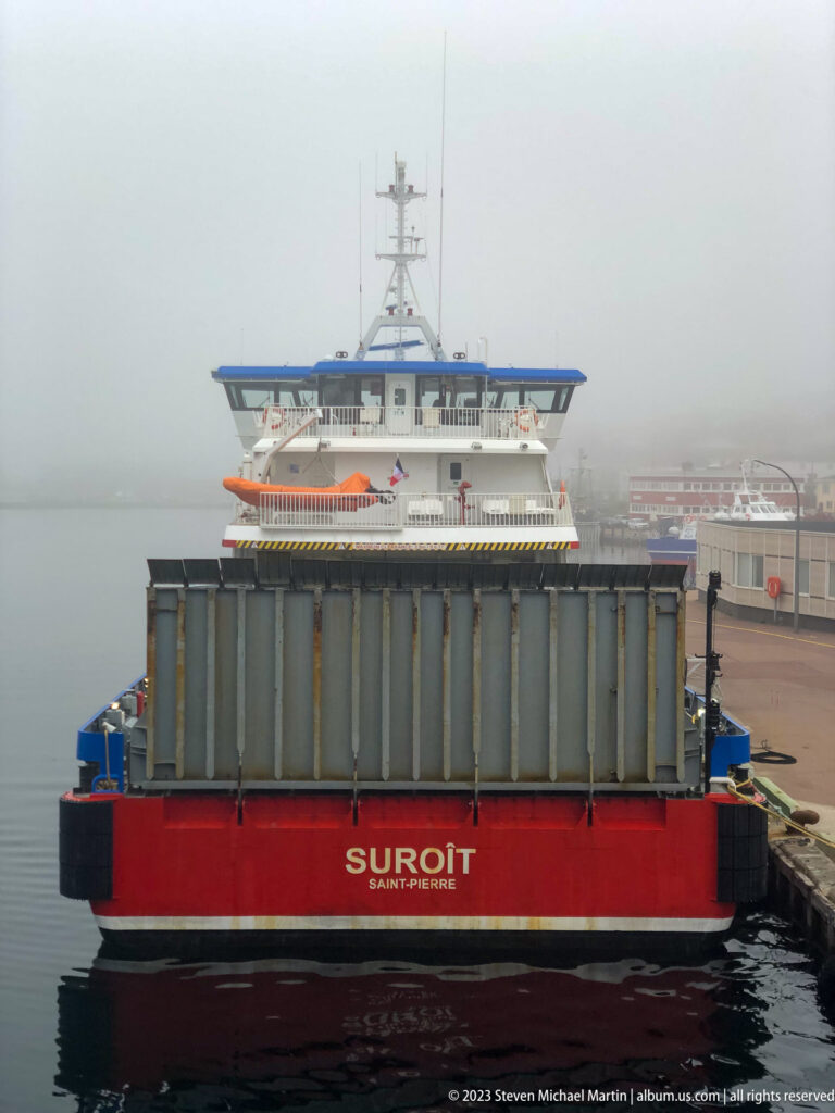 Red work barge named Suroît moored at Saint-Pierre, carrying large gray deck containers in fog.