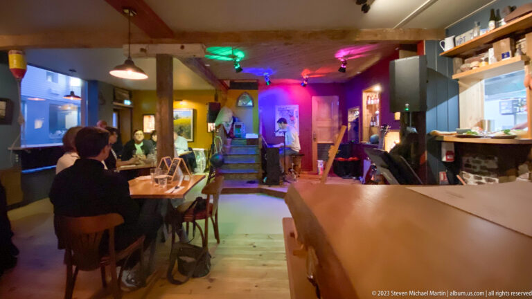 Long café-bar interior with seated patrons, wooden counter, colorful lighting, and a small music area at the rear.