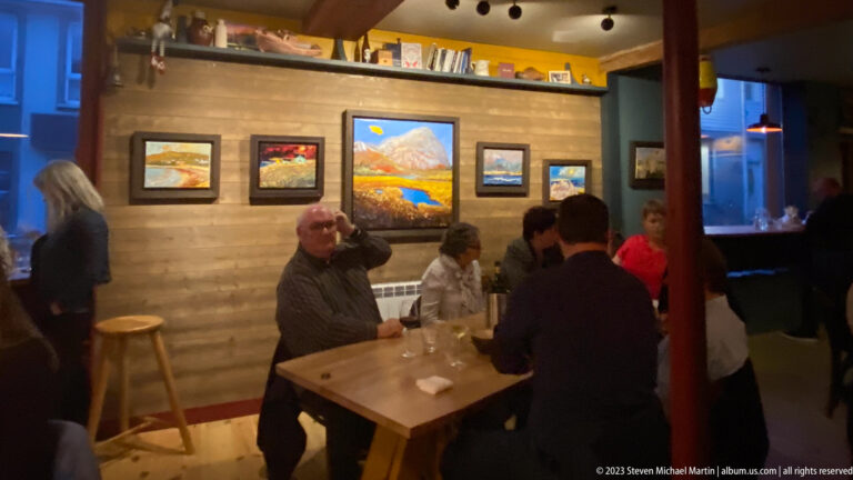 Patrons seated at a wooden table beneath several framed landscape paintings in a warmly lit dining room.