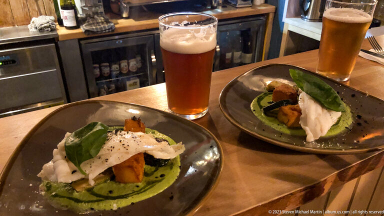 Two plated dishes with green sauce, roasted ingredients, white cheese or cream, and tall glasses of amber beer.