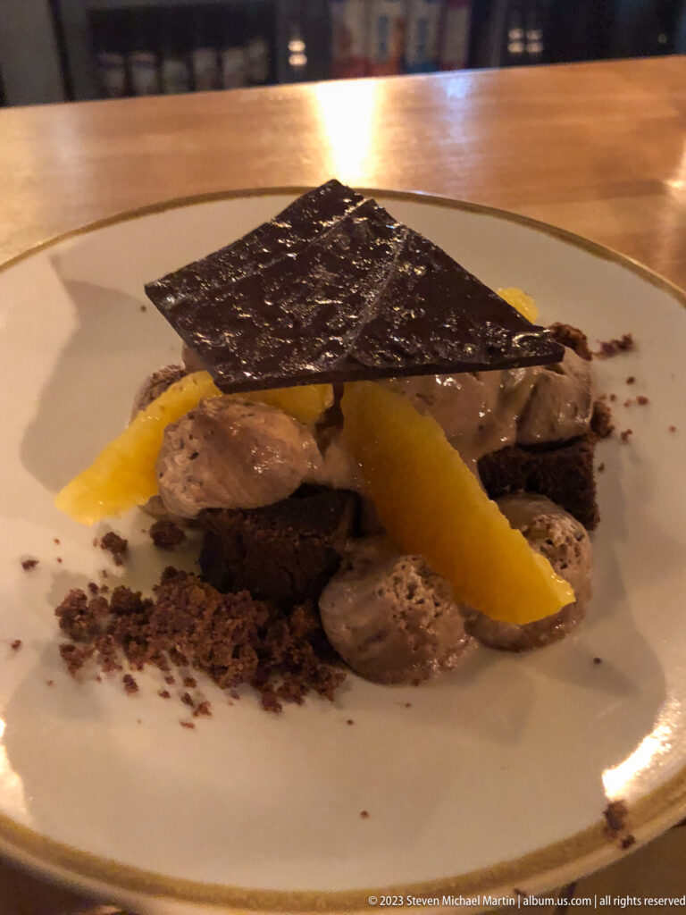 Large chocolate shard over a plated dessert with cream dollops, dark crumbs, and orange citrus segments.