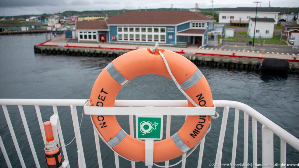 Orange lifebuoy marked Miquelon mounted on a white ferry rail, with a waterfront terminal beyond.