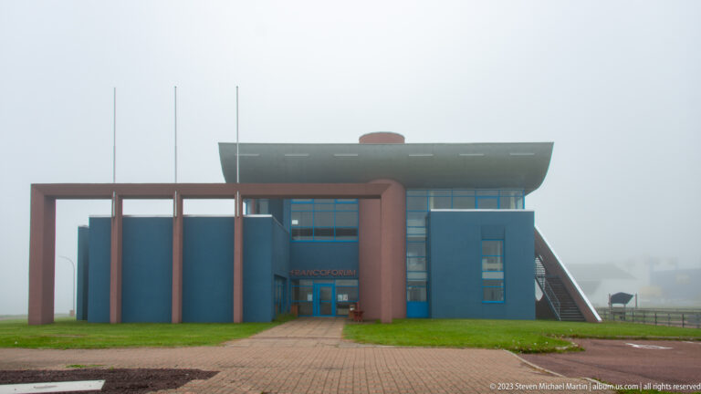 Modern blue public building with rose-colored frames, curved roof, tall windows, and exterior stairs in dense fog.