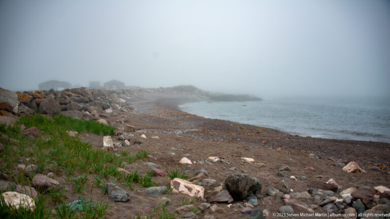 Dark gravel beach with scattered boulders and sparse plants curving into dense fog beside calm water.