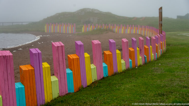 Bright pink, orange, yellow, turquoise, and green fence posts curving beside a foggy pebble beach.