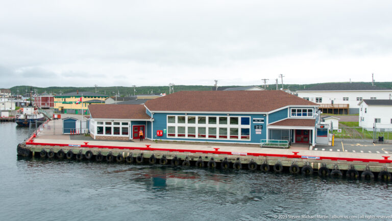 Blue-gray waterfront terminal with large windows on a quay, backed by small buildings under an overcast sky.