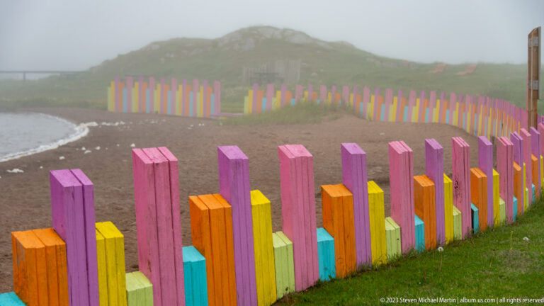 Close perspective of multicolored timber fence posts bending along a foggy shoreline.