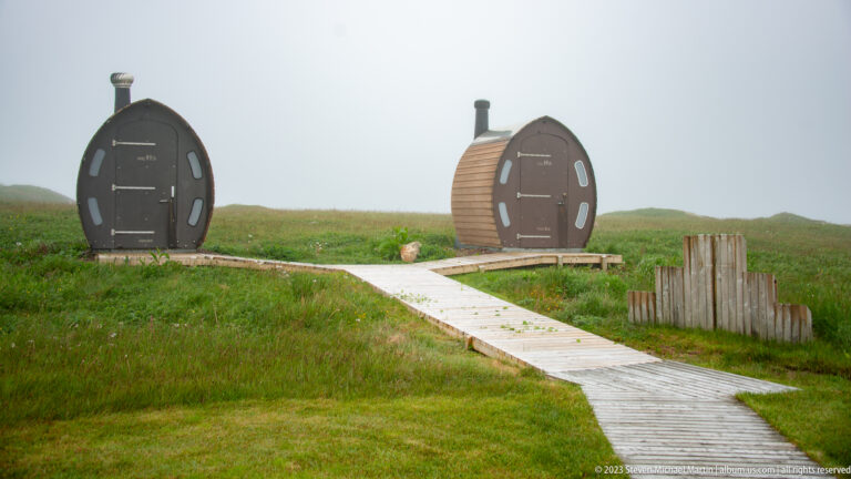 Two barrel-shaped wooden pod shelters with small windows and stovepipes beside a boardwalk in grass.