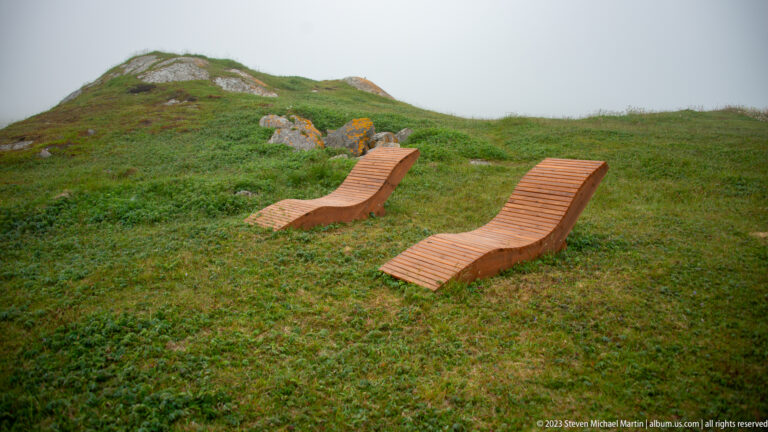 Two curved slatted wooden lounge chairs placed on a green hillside in fog.